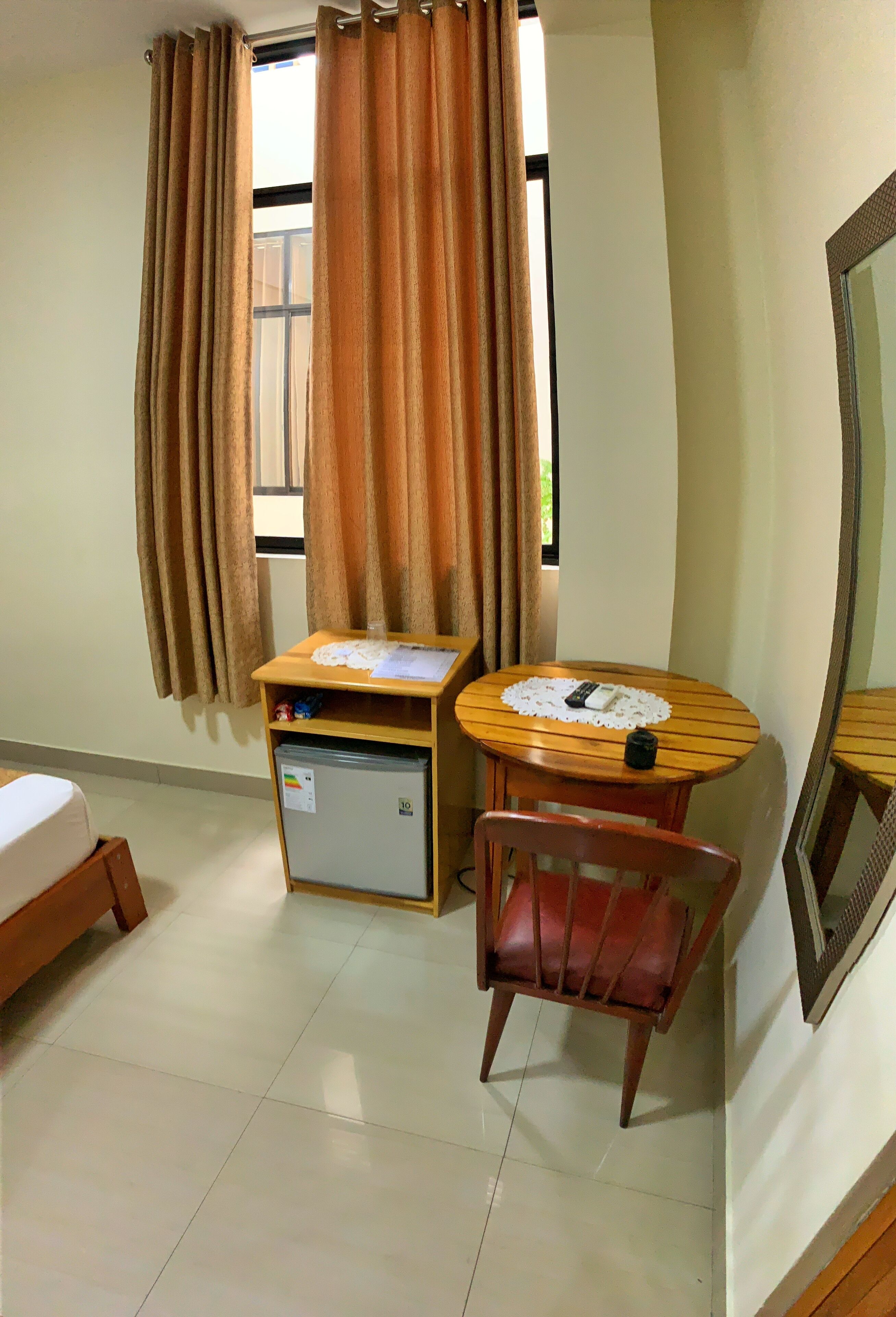 Habitación de hotel con una cama, una mesita con un control remoto, una silla, un mini refrigerador y una ventana con cortinas.