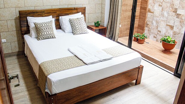 A neatly made bed with a wooden headboard, a small bedside table with a plant, and a view of a balcony with potted plants. - Studio 69 (Iquitos)