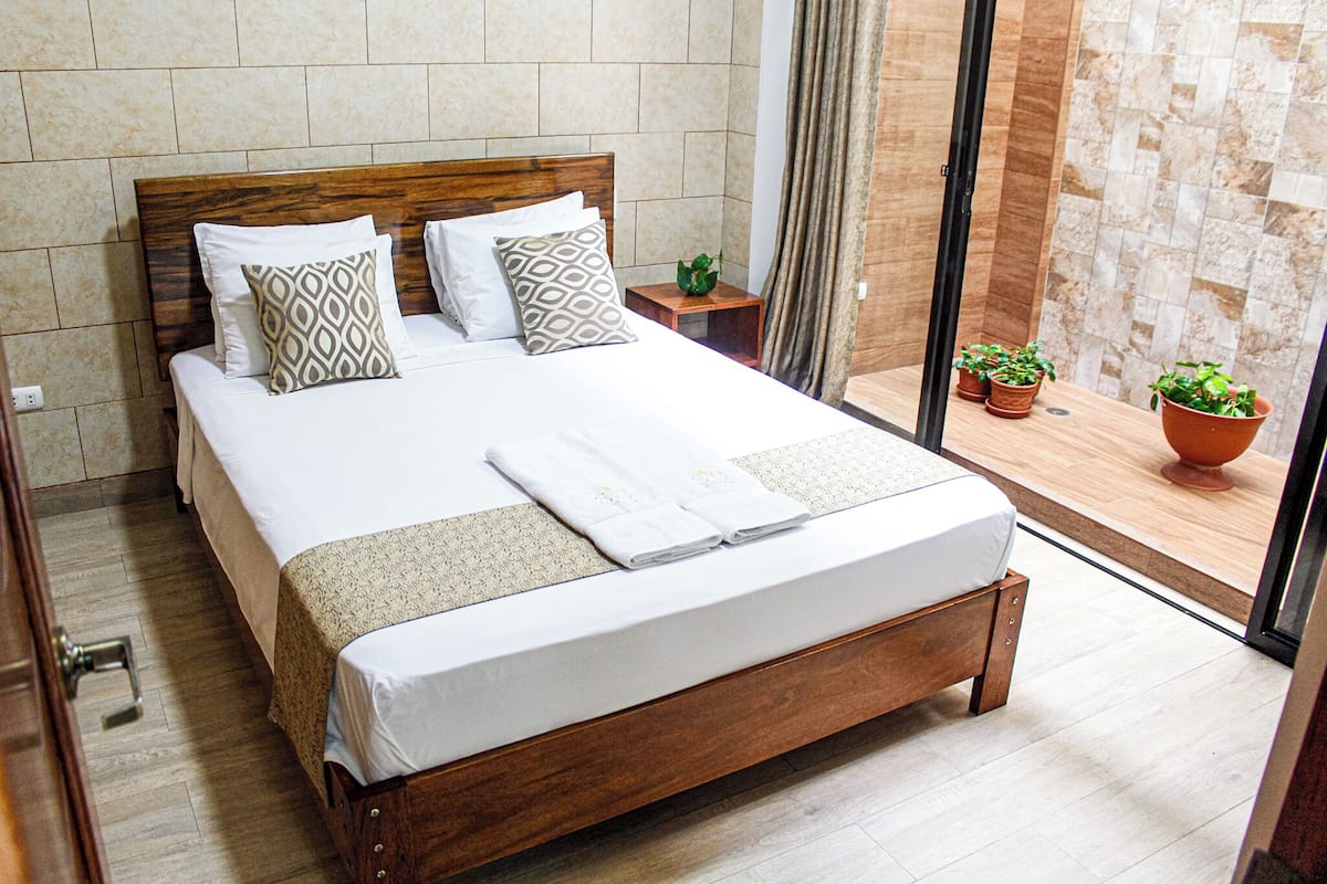 A neatly made bed with a wooden headboard, a small bedside table with a plant, and a view of a balcony with potted plants.