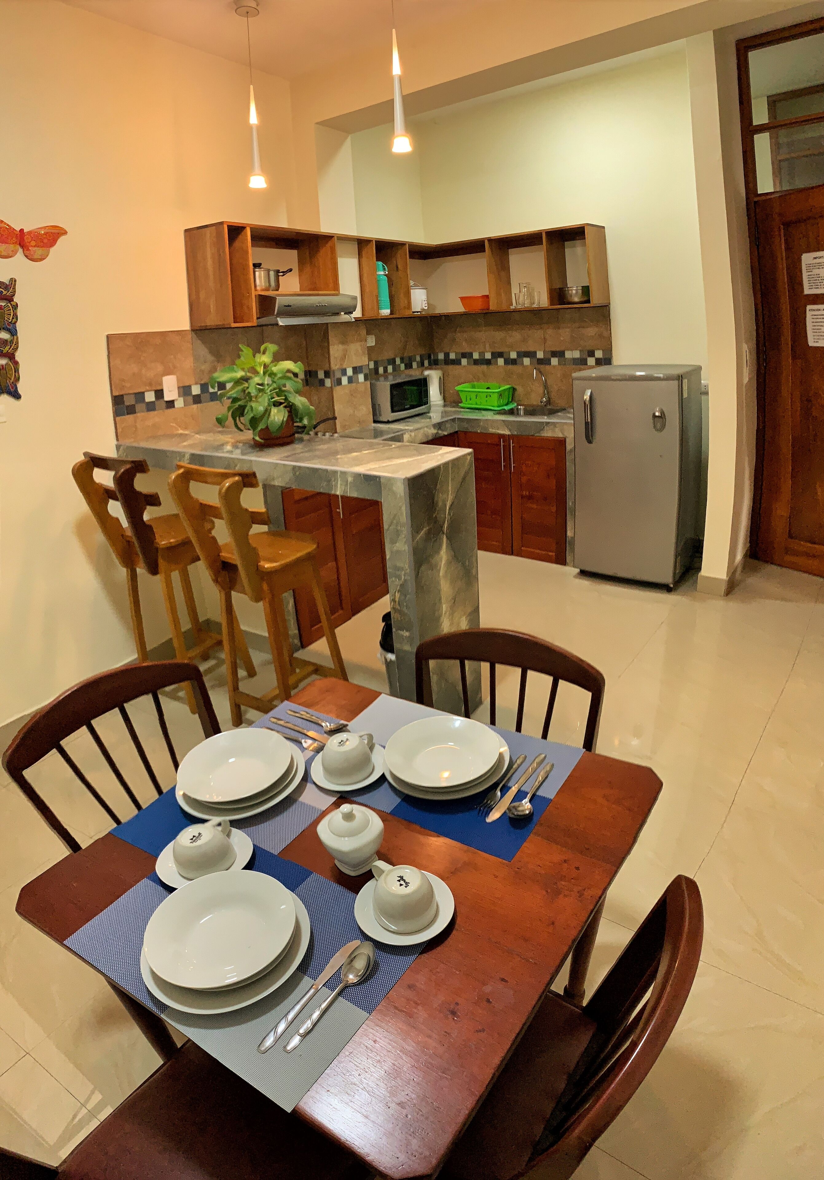 A dining area with a wooden table set for four, a kitchen in the background with modern appliances, and a decorative plant on the counter.
