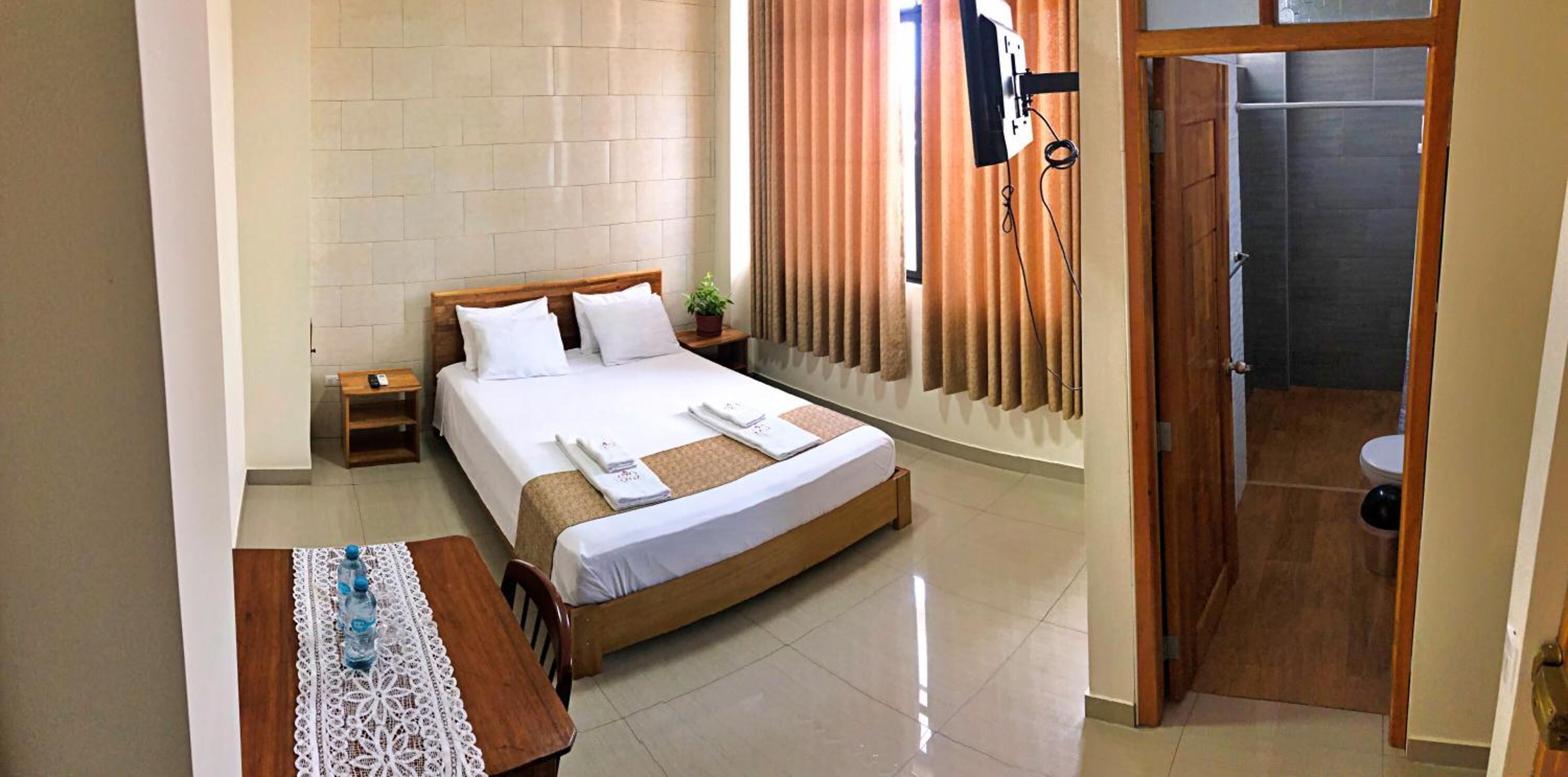 A hotel room with a large bed, a wooden nightstand, a table with a lace tablecloth, a potted plant, and a view of a shower area.