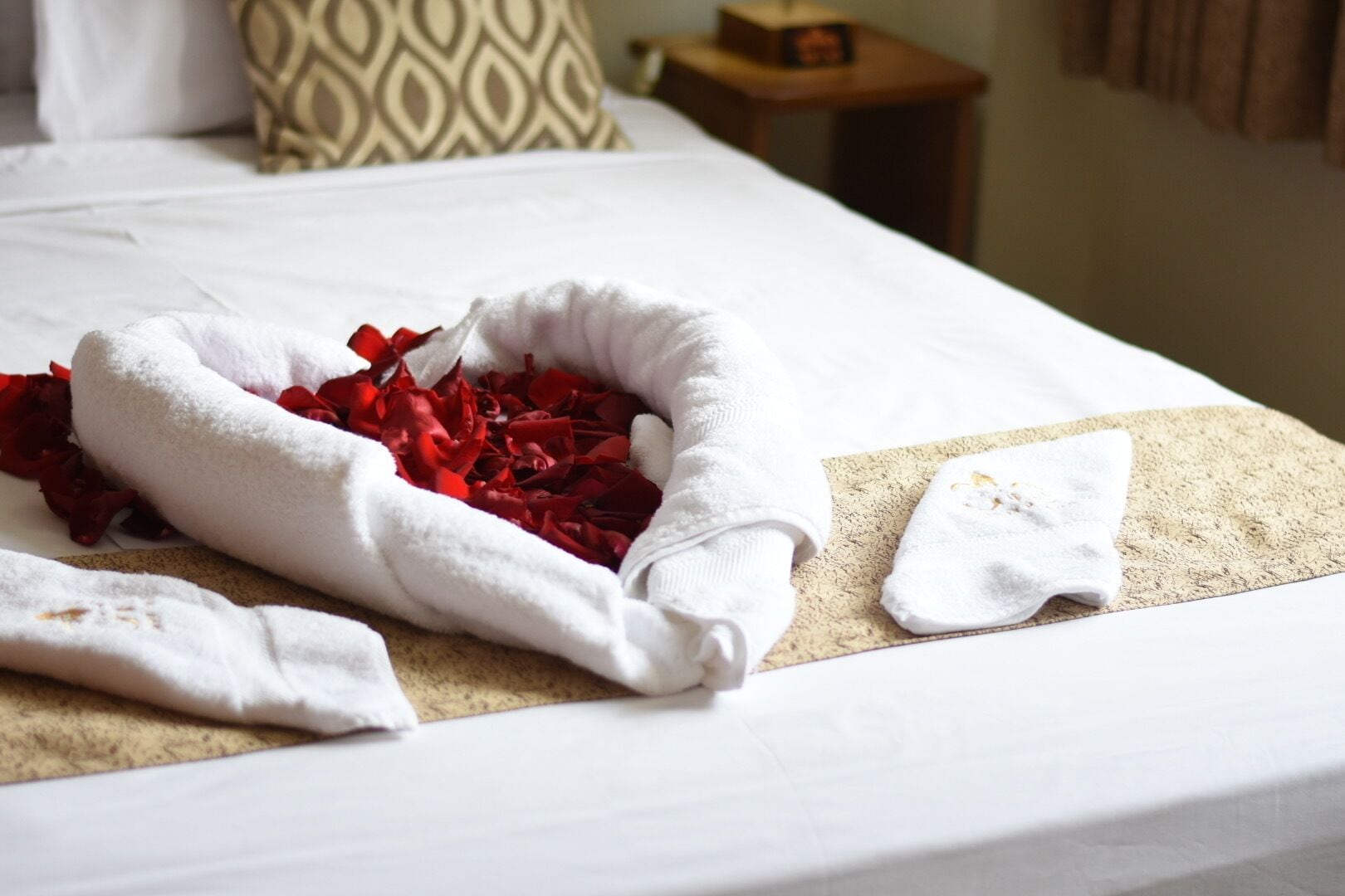 A neatly made bed with a white towel, a white bathrobe, and a basket filled with red rose petals.
