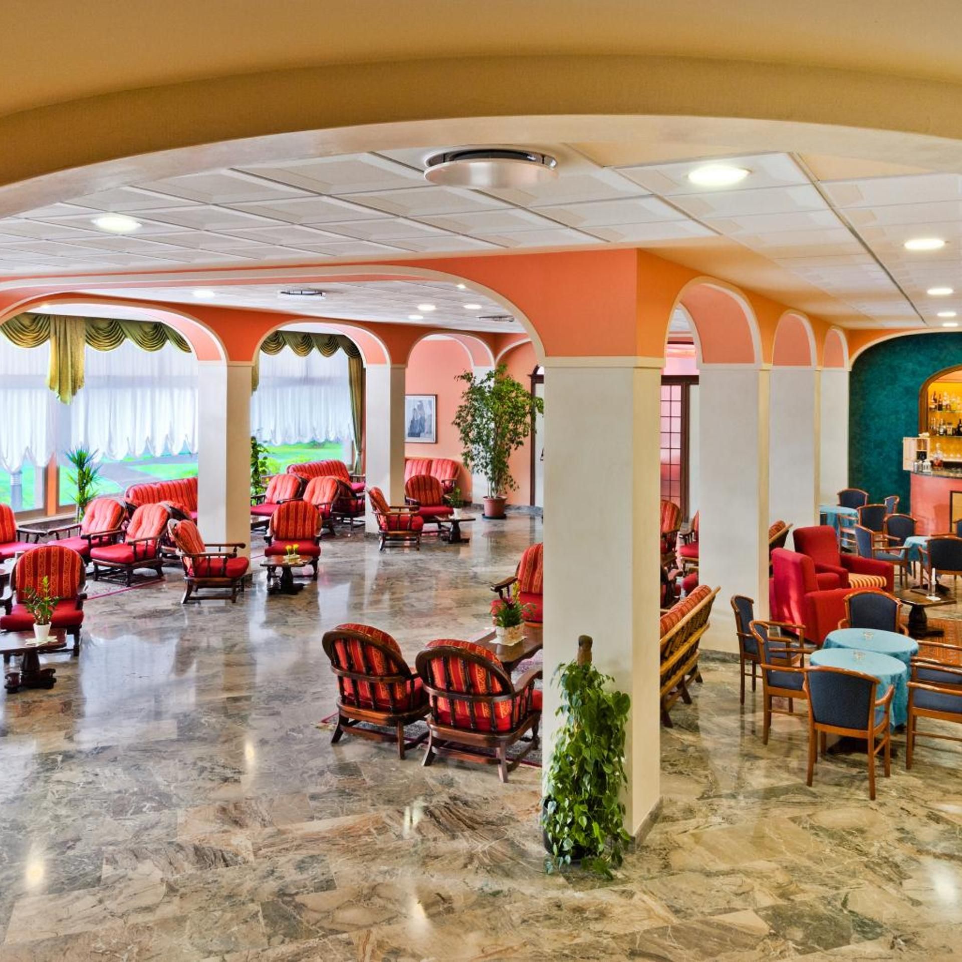 A spacious lobby with red upholstered chairs, a bar area, and a view of a green landscape through the windows.