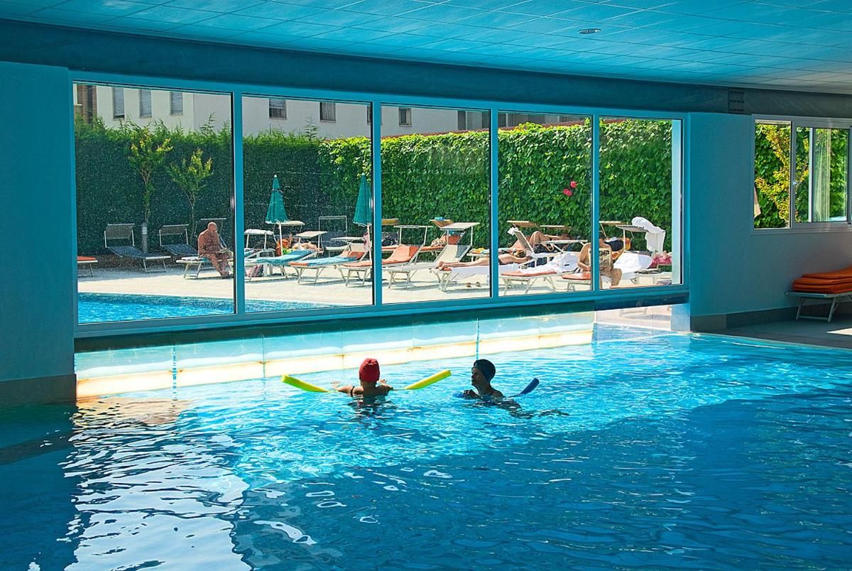 An indoor swimming pool with clear glass walls, a view of the outside, and lounge chairs.