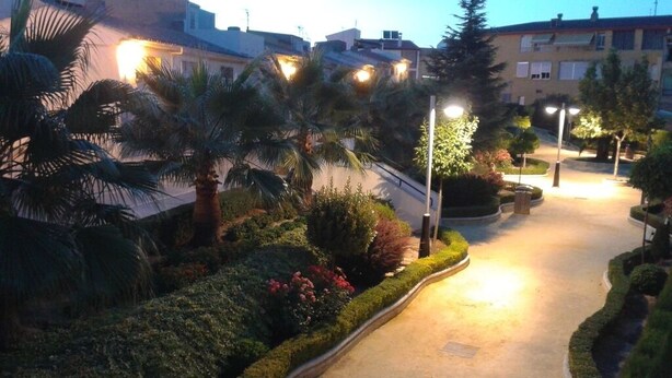 Un sendero bien iluminado con palmeras y arbustos, que conduce a un edificio con múltiples ventanas y balcones.