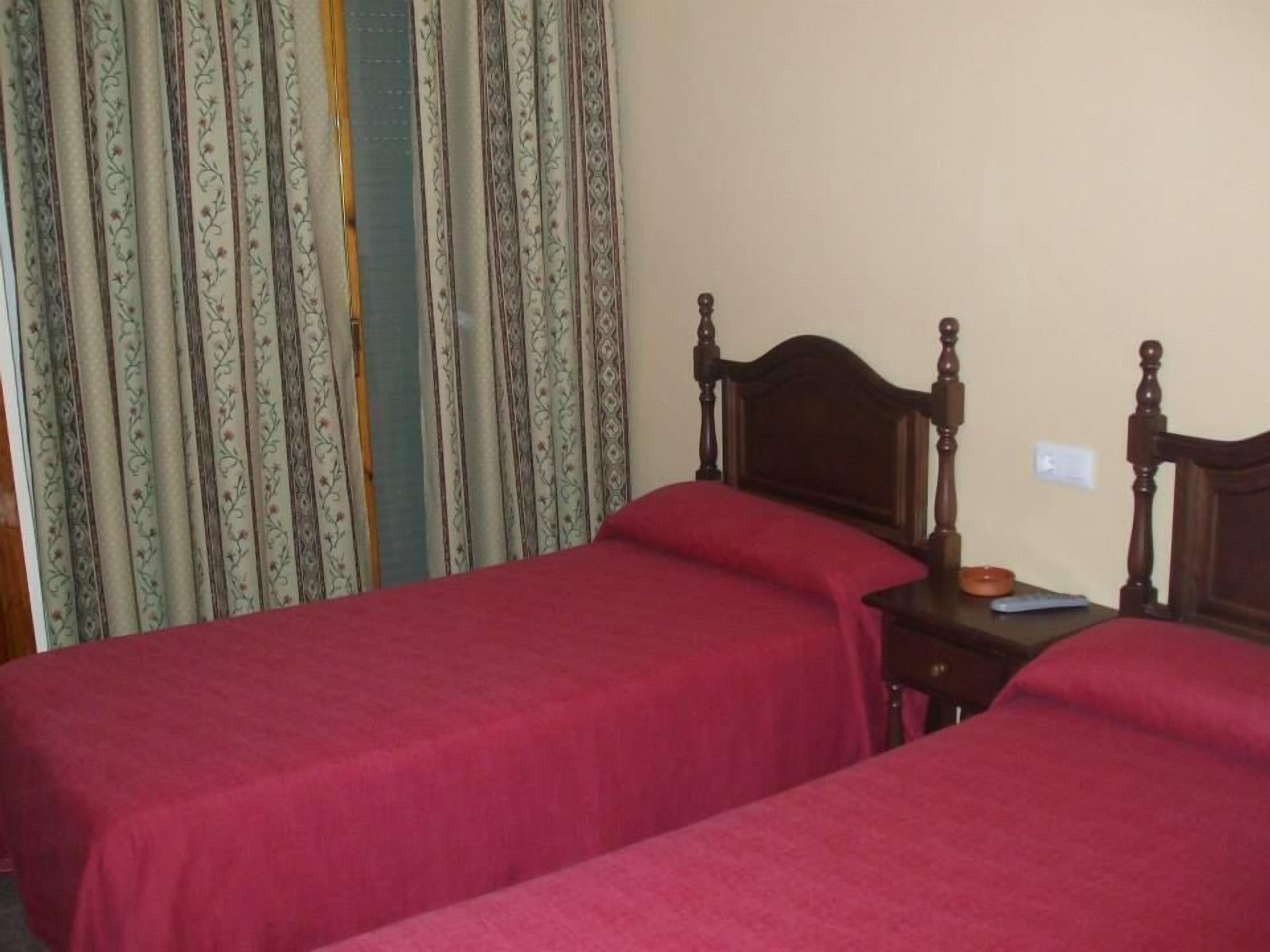 Una habitación con dos camas individuales, cabecera de madera, mesita de noche pequeña con lámpara y cortina estampada con motivos florales.