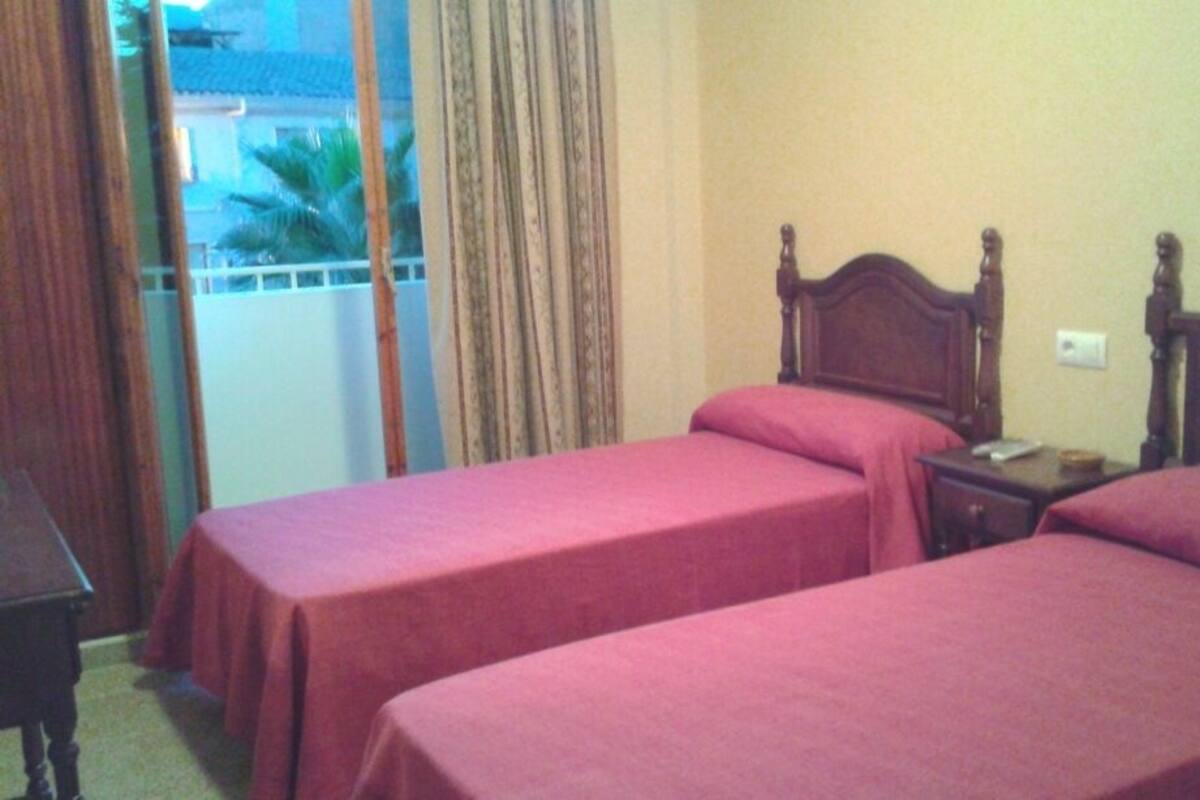 A hotel room with two single beds, a wooden headboard, a nightstand, and a view of a balcony with curtains.