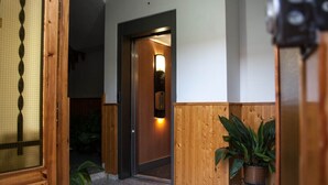Un pasillo interior con una puerta de madera que da a una habitación, una planta en maceta y un aplique en la pared.