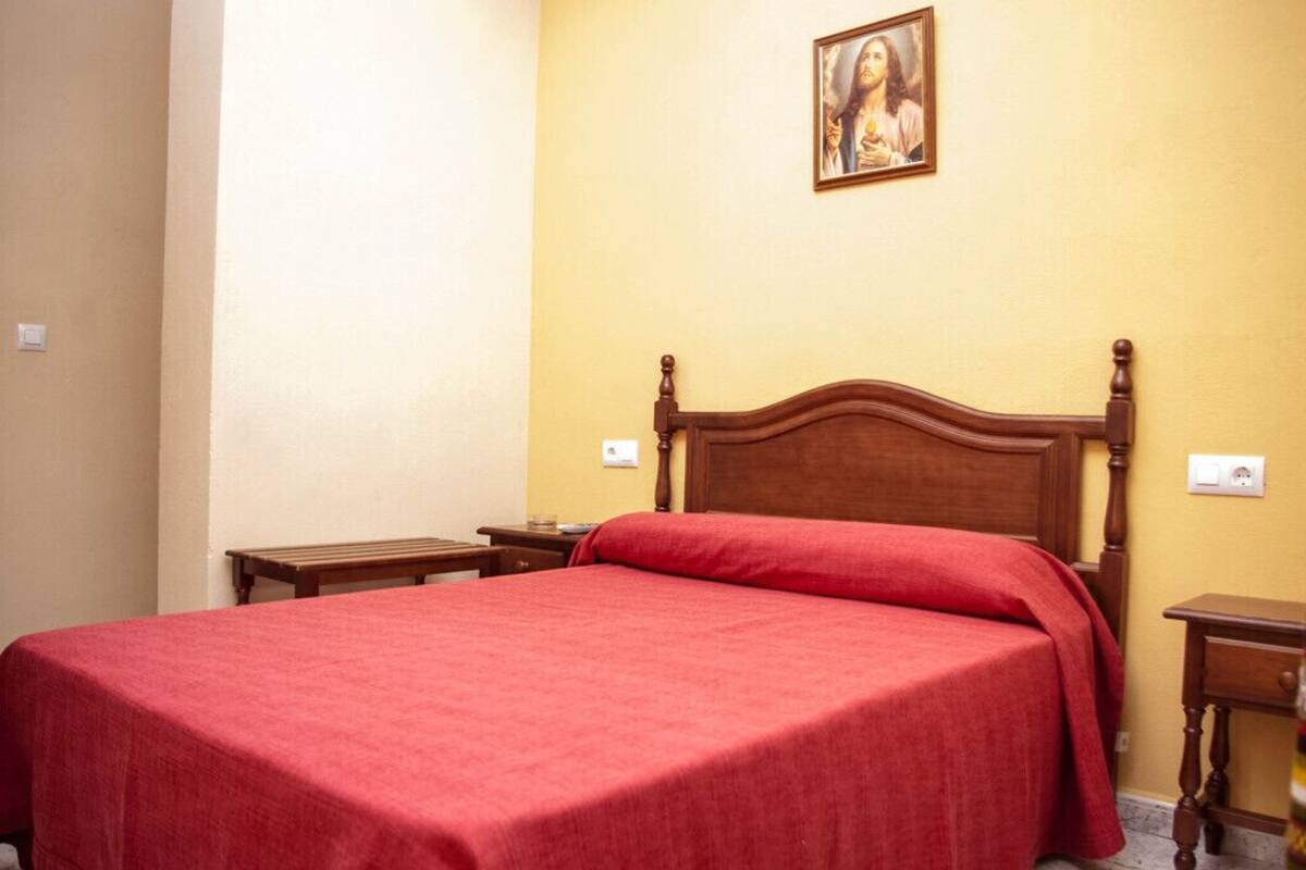 A neatly made bed with a wooden headboard and a red bedspread in a room with yellow walls and a small painting above the bed.