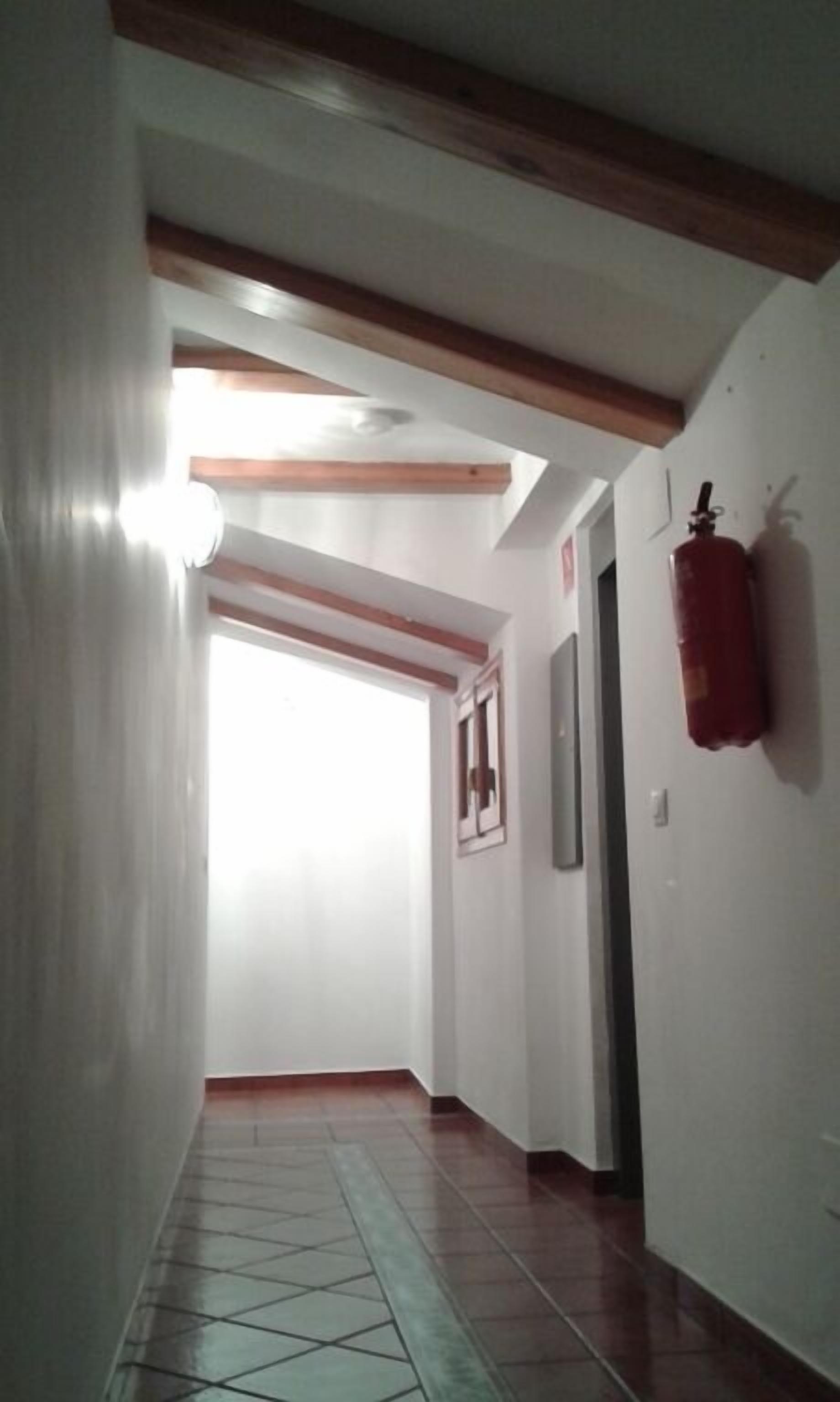 Un couloir doté d’un sol carrelé, de poutres en bois au plafond, d’un extincteur fixé au mur et d’une porte au fond.