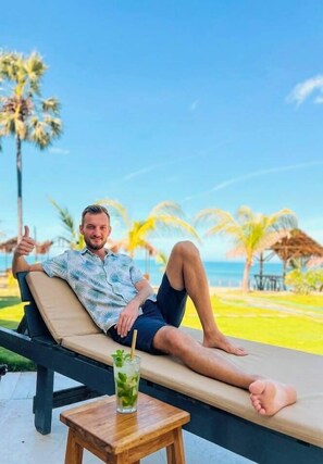 A man relaxing on a lounge chair by a pool with a drink on a small table, palm trees, and a clear sky in the background. - Guitar Bay Arugambay (Arugam Bay)