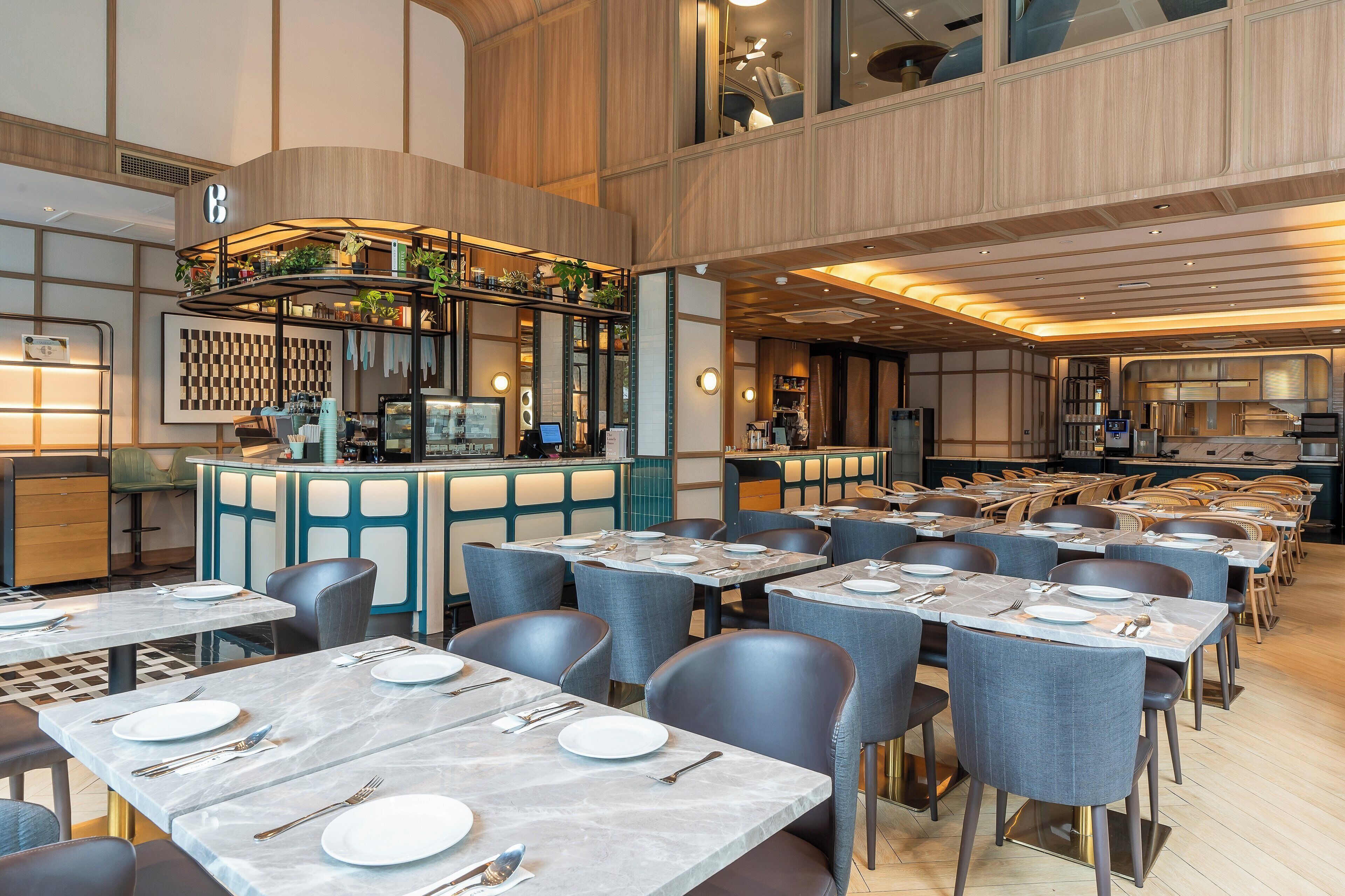 A modern restaurant with marble-topped tables, grey upholstered chairs, and a central bar area with plants and a computer monitor.