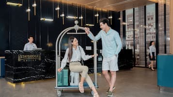 A reception area with a black marble counter labeled "Staybridge Suites," a woman seated on a luggage cart, and a man standing beside her.