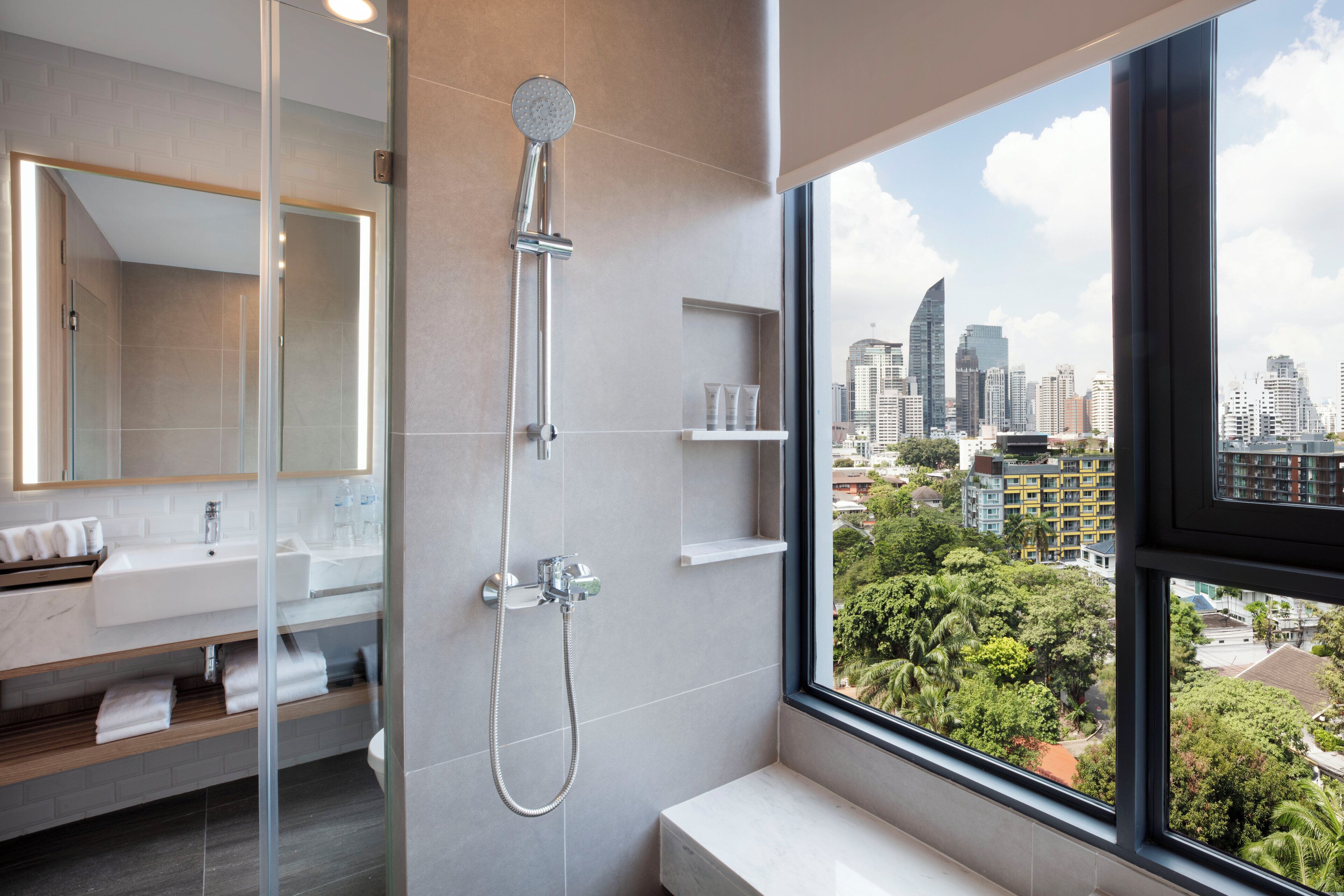 A modern bathroom with a glass shower enclosure, a large mirror, a sink with a marble countertop, and a window offering a city view.