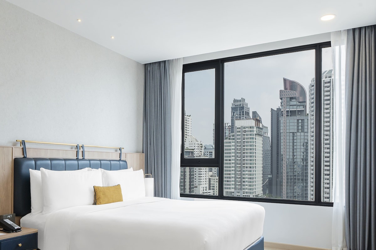Une chambre d’hôtel moderne avec un grand lit, une table de chevet et une vue sur les grands immeubles, visible à travers une grande fenêtre.