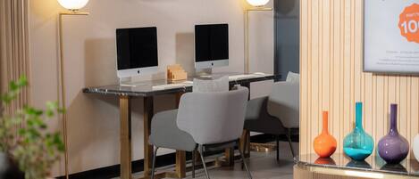 A modern office space with two desks, each with a computer monitor, a lamp, and a chair. There are colorful vases on a shelf and a framed poster on the wall.