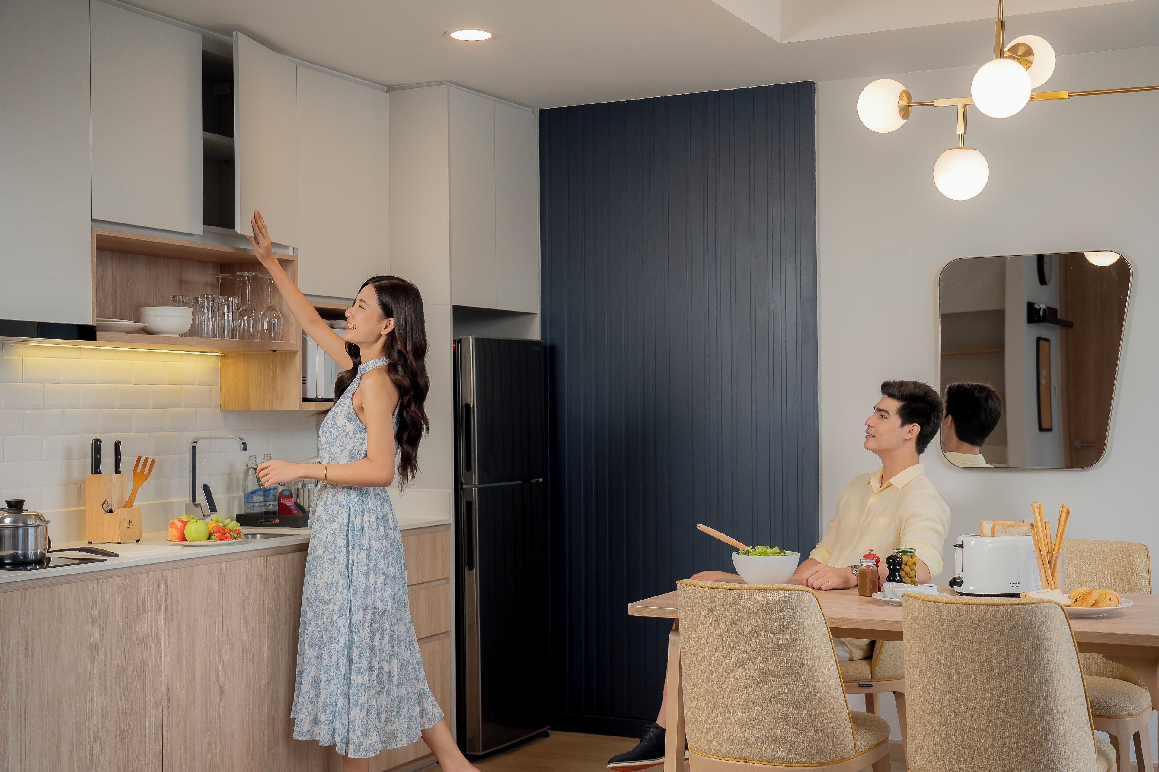 A modern kitchen with a woman reaching for a shelf, a man dining at a table, and a dining area with chairs and a table.