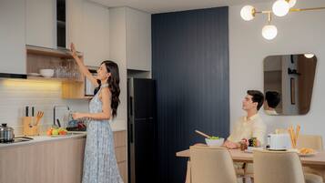 A modern kitchen with a woman reaching for a shelf, a man dining at a table, and a dining area with chairs and a table.