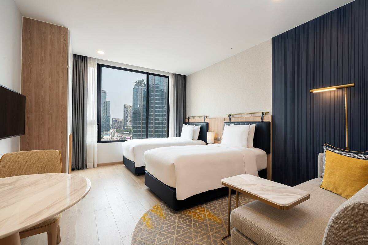 Une chambre d’hôtel avec deux lits, une table à manger, un canapé et une grande fenêtre donnant sur la ville.