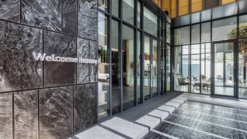A modern hotel entrance with a 'Welcome home' sign, large glass doors, and a view of the outdoor patio area.