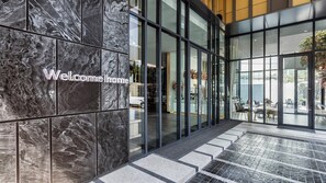 A modern hotel entrance with a 'Welcome home' sign, large glass doors, and a view of the outdoor patio area.