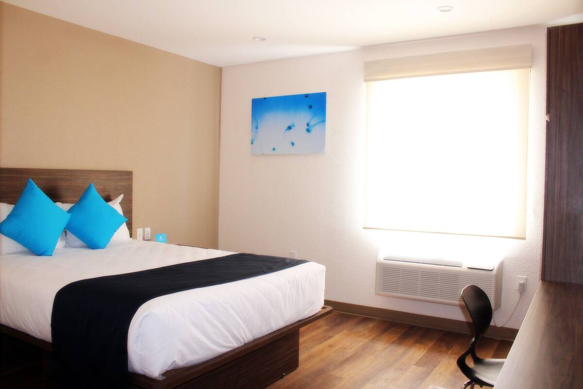 A hotel room with a large bed, a window with blinds, a wall-mounted air conditioner, and a painting on the wall.