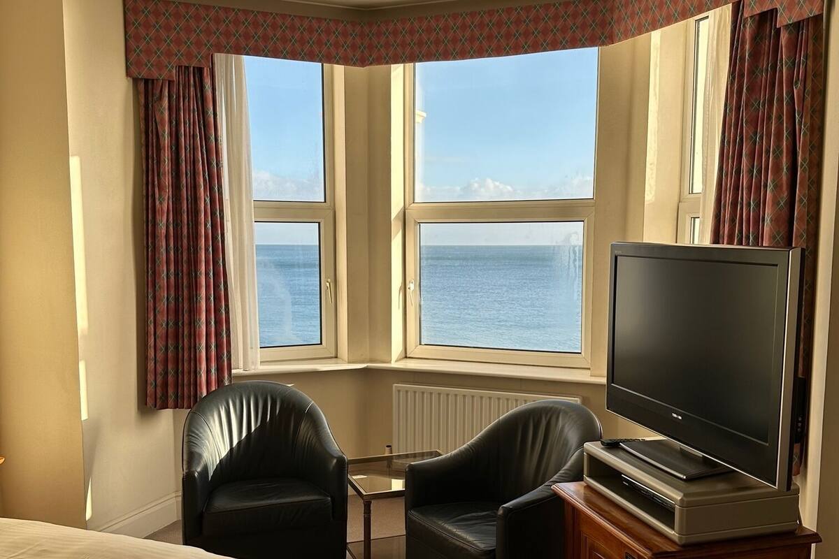 A hotel room with a large bed, two black leather chairs, a small table, a television on a wooden stand, and a view of the sea through two windows.