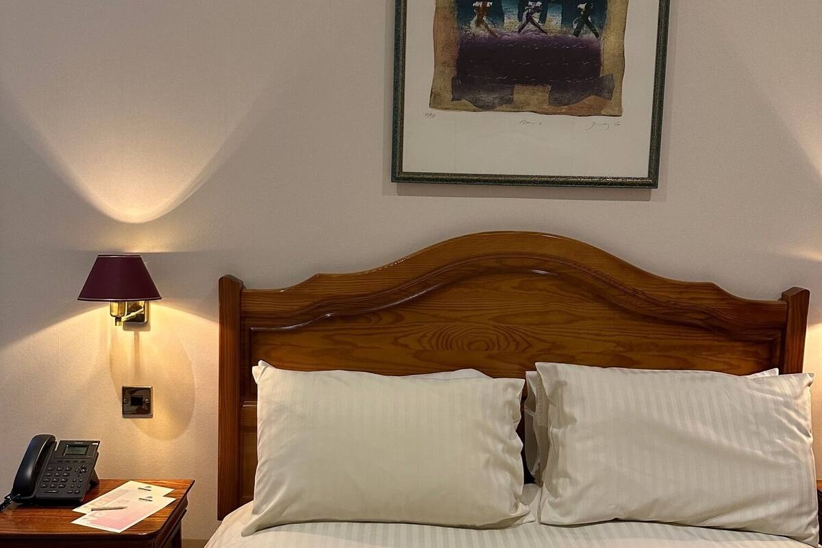 A hotel room with a wooden headboard, two white pillows, a bedside table with a lamp, a phone, and a framed artwork on the wall.