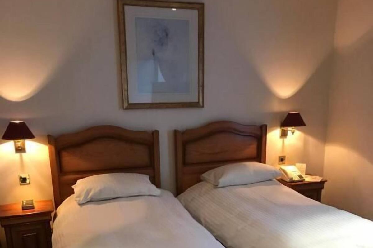 A hotel room with two single beds, each with a wooden headboard, a bedside table, and a lamp. There is a framed picture on the wall above the beds.