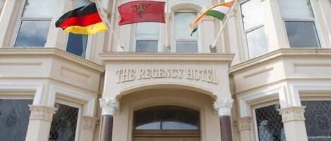 Un edificio imponente con una entrada destacada, banderas y el nombre "The Regency Hotel" sobre el acceso.