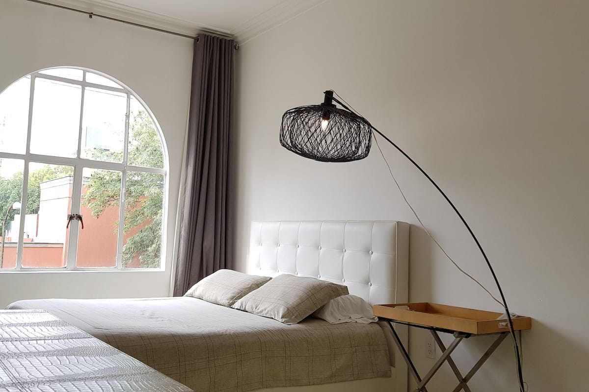 A bedroom with a large window, a bed with a white headboard, a bedside table, and a hanging light fixture.