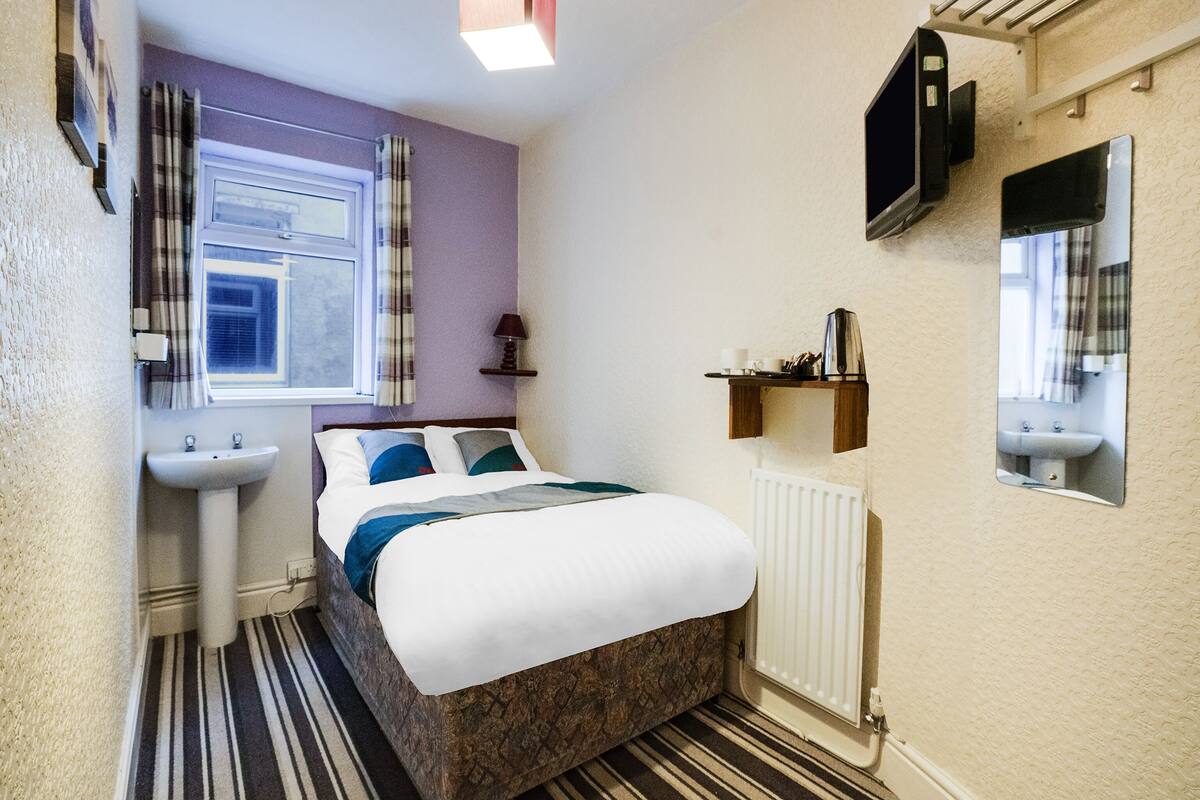 Habitación de hotel pequeña con cama, ventana con cortinas, mesita de noche con lámpara, estante con artículos de aseo y radiador.