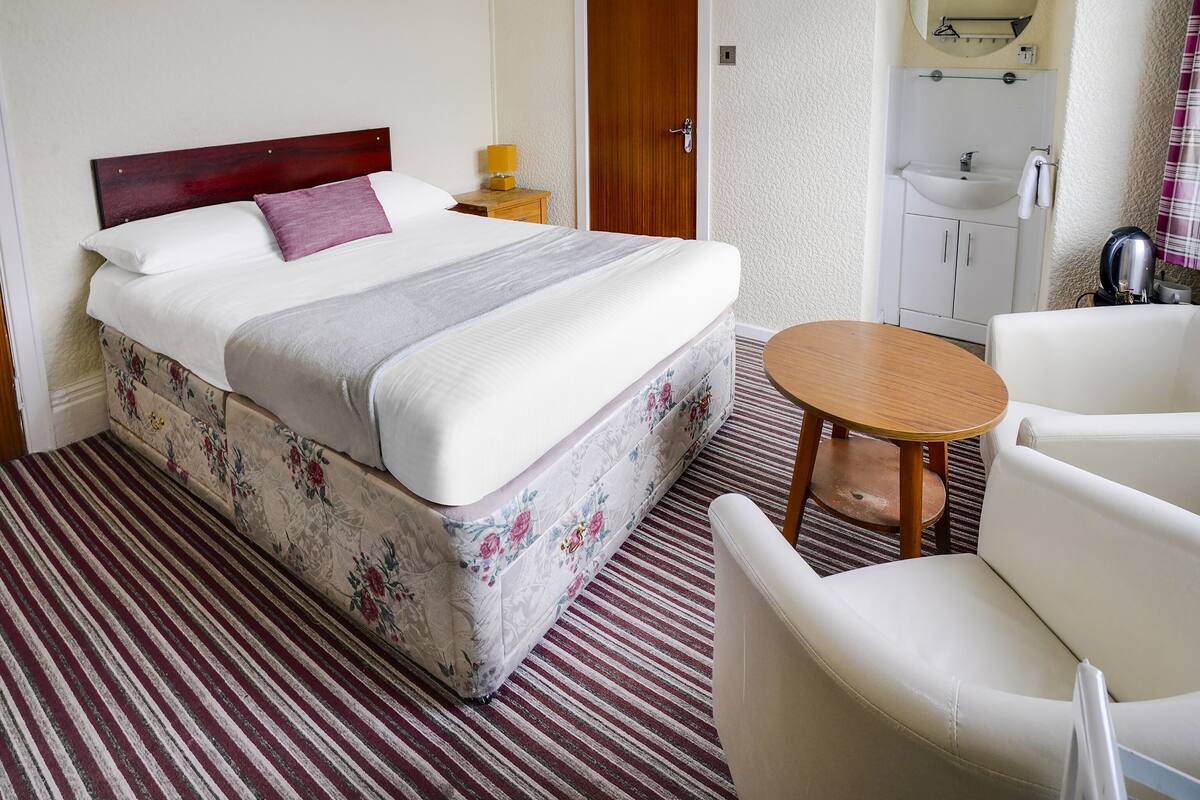 Una habitación de hotel con cama, una mesita redonda pequeña, una silla, una mesita de noche, un espejo y un baño con lavabo e inodoro.