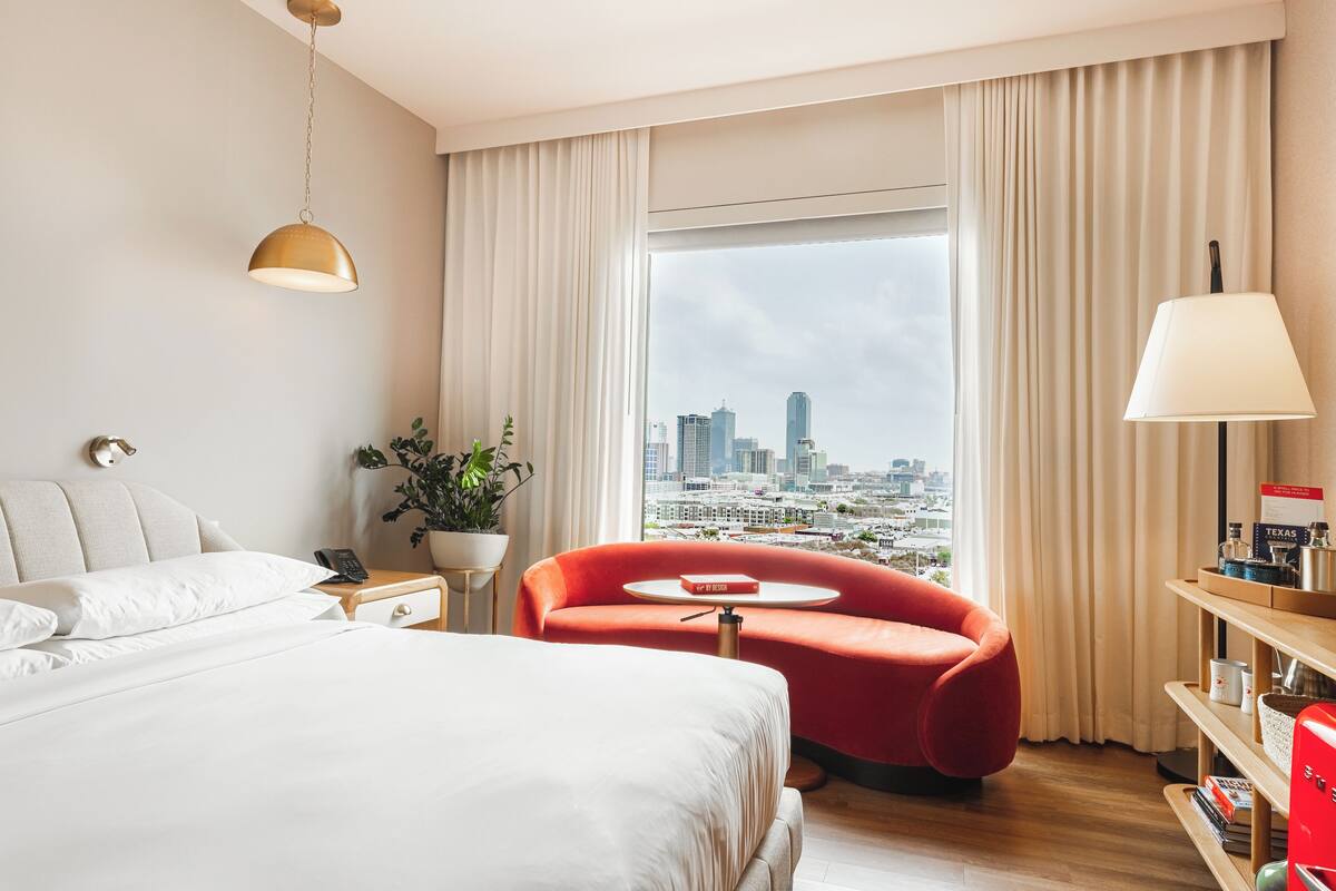 Hotelkamer met een bed, een rode fauteuil, een klein tafeltje, een lamp, een nachtkastje, een potplant en uitzicht op een stadsgezicht door grote ramen.