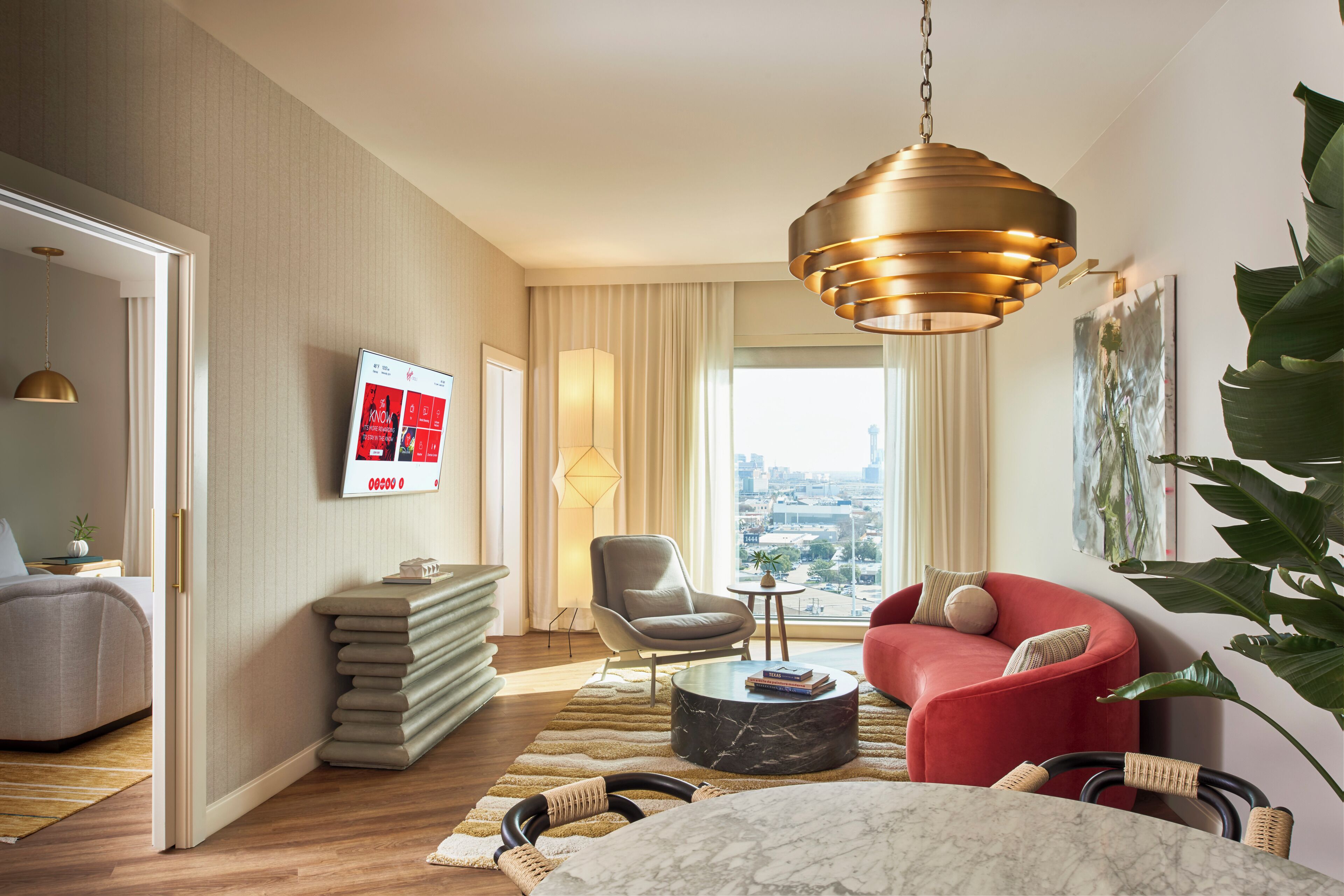 A modern hotel room with a large window, a red sofa, a grey armchair, a marble coffee table, and a hanging gold pendant light.