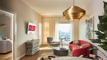 A modern hotel room with a large window, a red sofa, a grey armchair, a marble coffee table, and a hanging gold pendant light.
