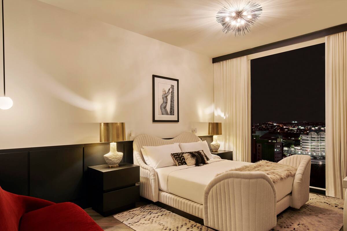 Een moderne slaapkamer met een groot bed, twee nachtkastjes met lampen, een stadszicht door een groot raam, en een rode stoel.