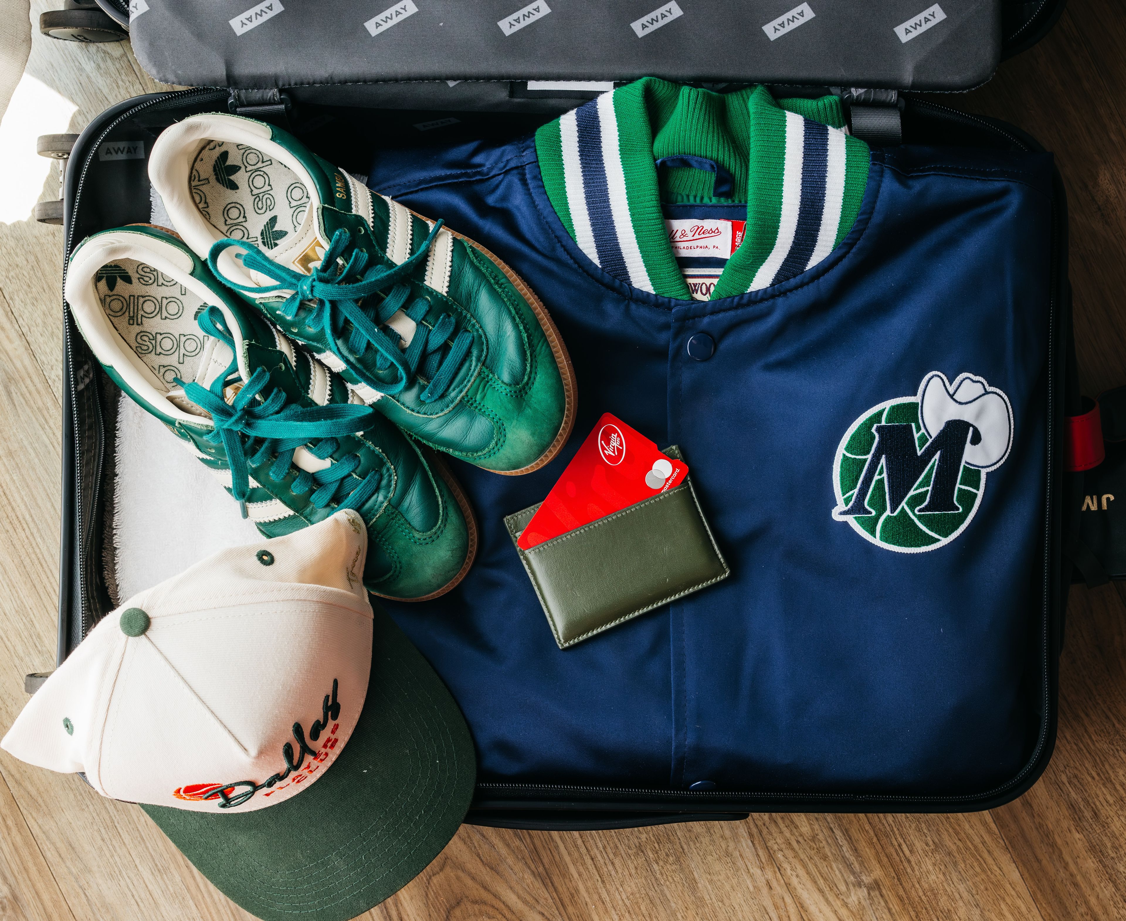 A suitcase containing a pair of green sneakers, a navy blue jersey with a logo, a red credit card, and a cap.
