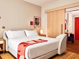 A hotel room with a large bed, a bedside table with a lamp, a red door, and a view of the hallway.
