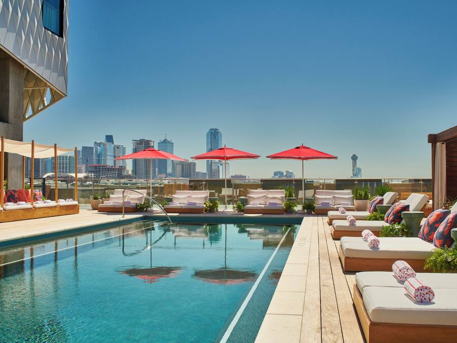 A rooftop pool with lounge chairs and red umbrellas, a city skyline in the background, and a clear sky.