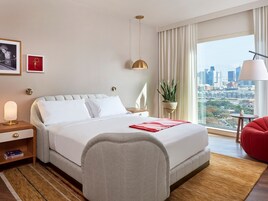 Una habitación de hotel con una cama grande, una mesita de noche con lámpara, una silla roja, una planta en maceta y vistas a un horizonte urbano.
