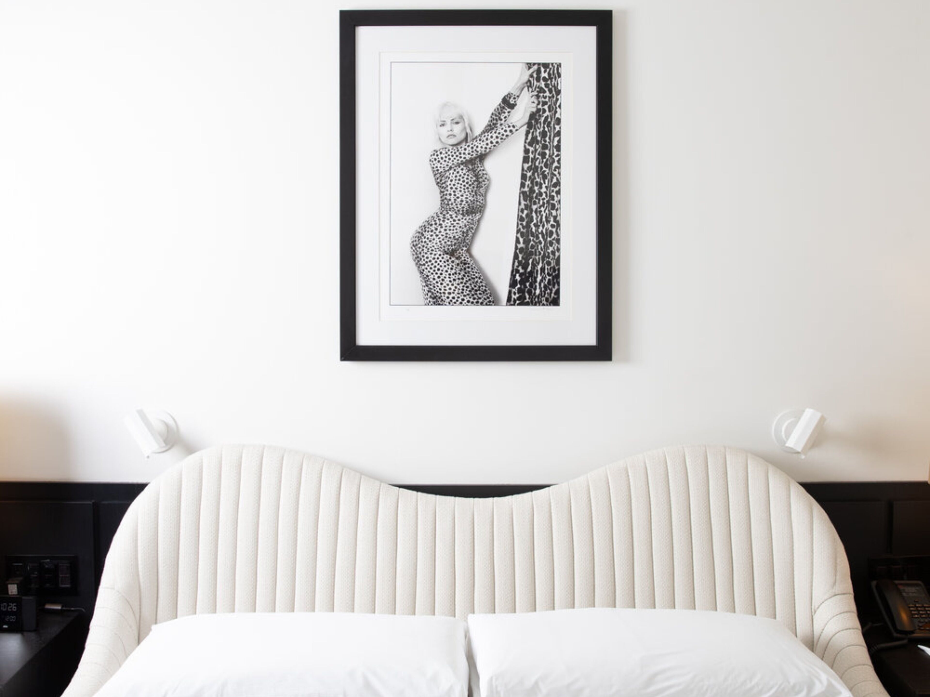 Fotografía en blanco y negro enmarcada de una persona con un vestido estampado colgada en la pared sobre una cama con cabecera acanalada.