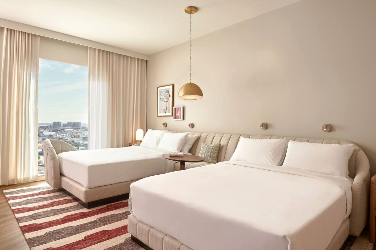 Een hotelkamer met twee bedden, een groot raam met gordijnen, uitzicht op de stad en een moderne kroonluchter.