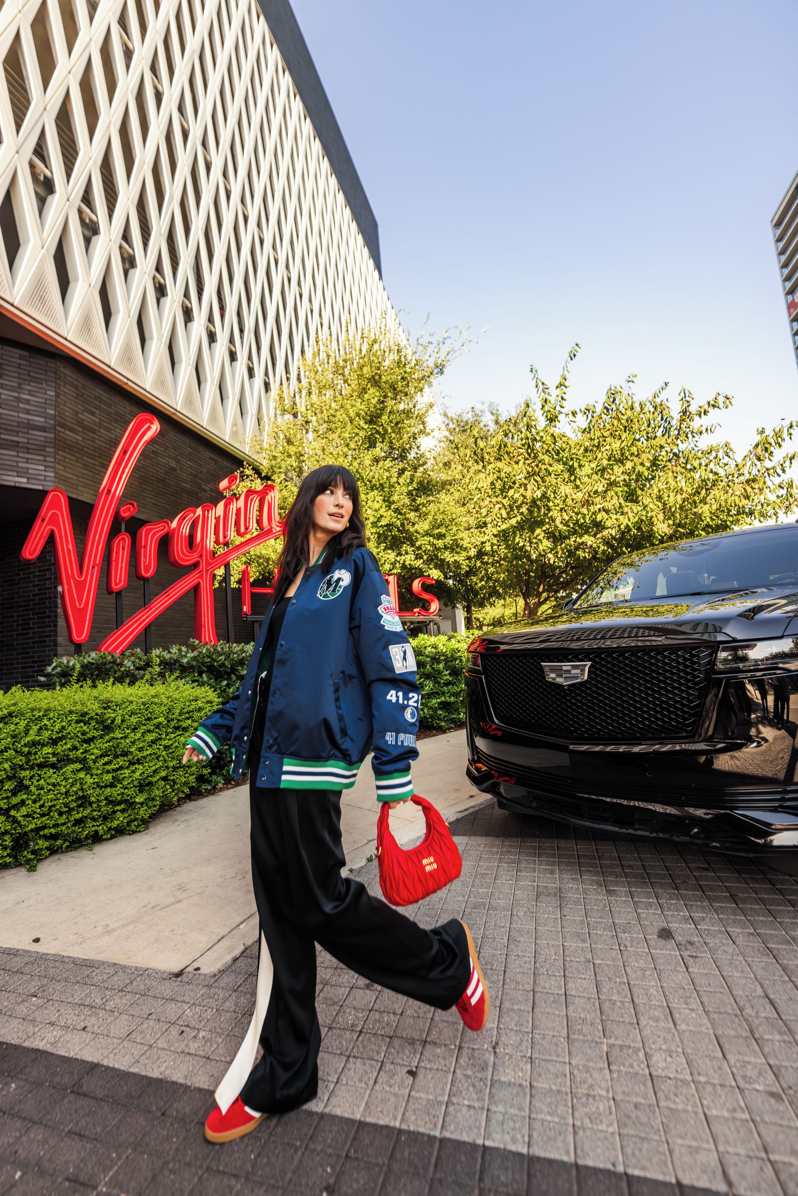 A person walking on a paved area with a building featuring a distinctive geometric facade and a neon sign in the background.