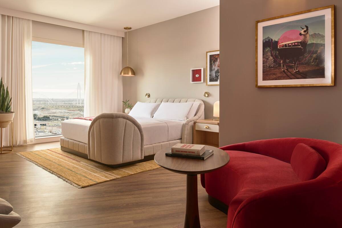 Een moderne hotelkamer met een bed, een rode fauteuil, een klein tafeltje en uitzicht op de stad door grote ramen.