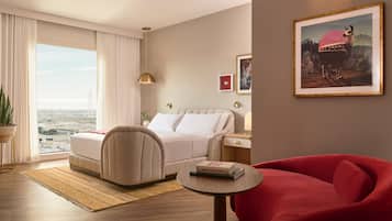 Ein modernes Hotelzimmer mit Bett, einem roten Sessel, einem kleinen Tisch und Blick auf die Stadt durch große Fenster.