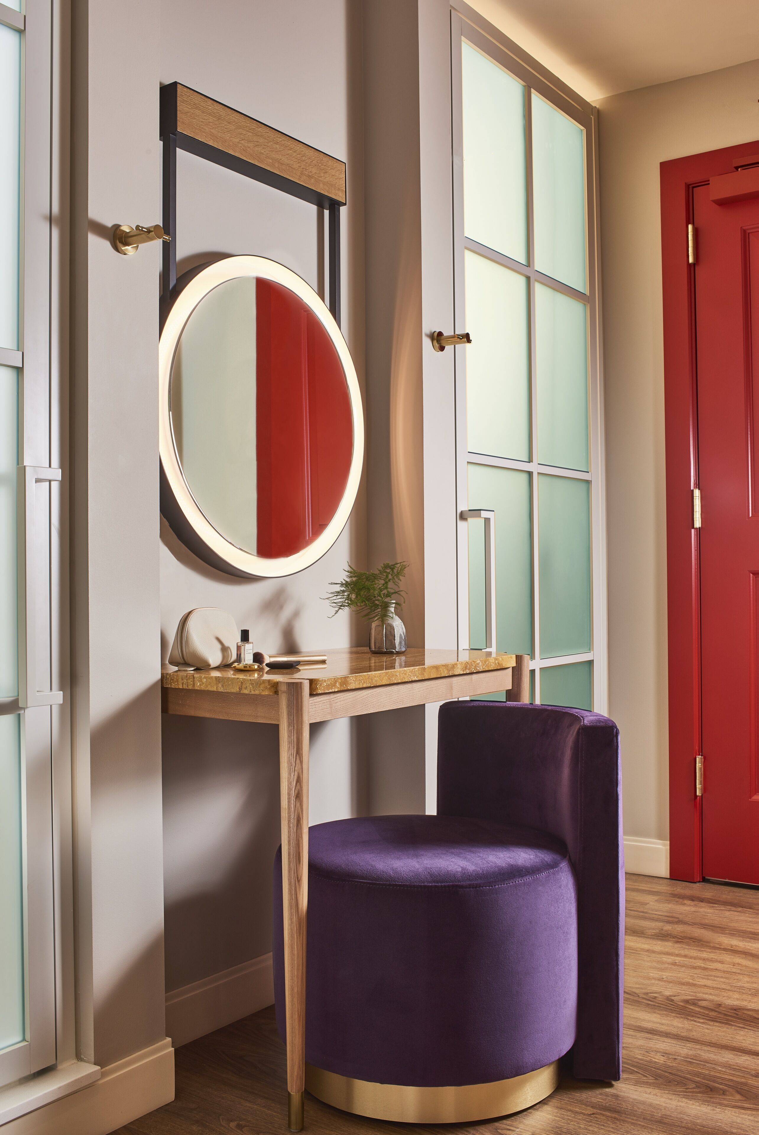Una habitación con un espejo redondo, un tocador de madera, una silla morada y una puerta roja.
