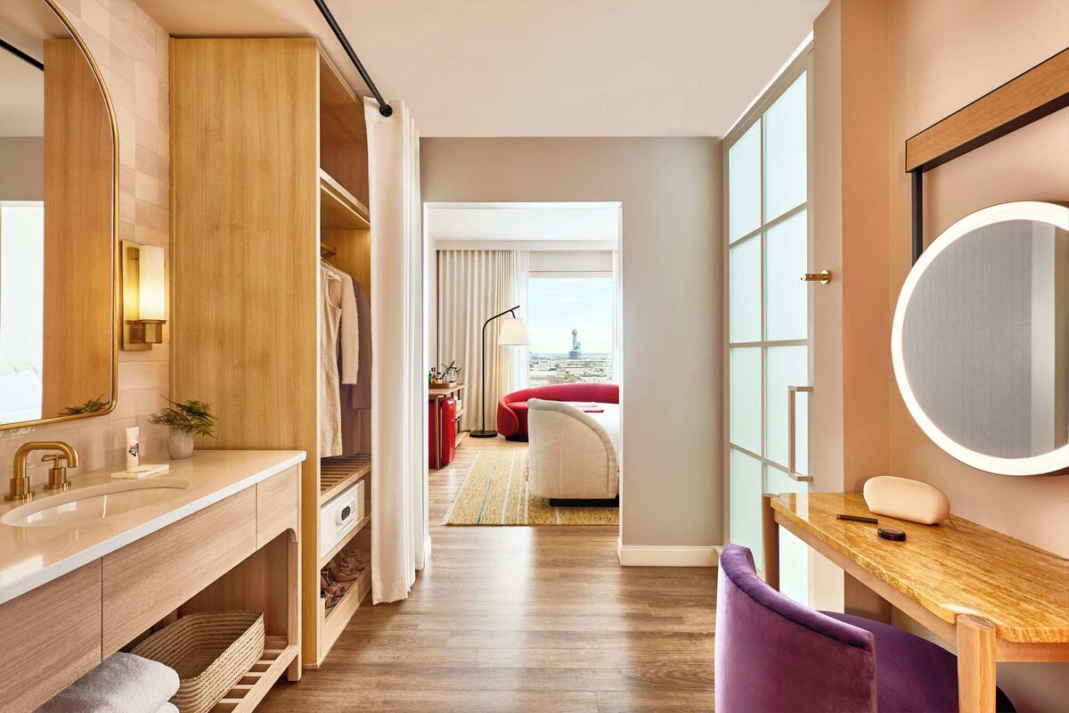 Een moderne hotelkamer met een houten wastafel, een ronde spiegel, een paarse stoel en uitzicht op de stad door grote ramen.