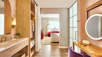 A modern hotel room with a wooden vanity, a round mirror, a purple chair, and a view of the city through large windows.