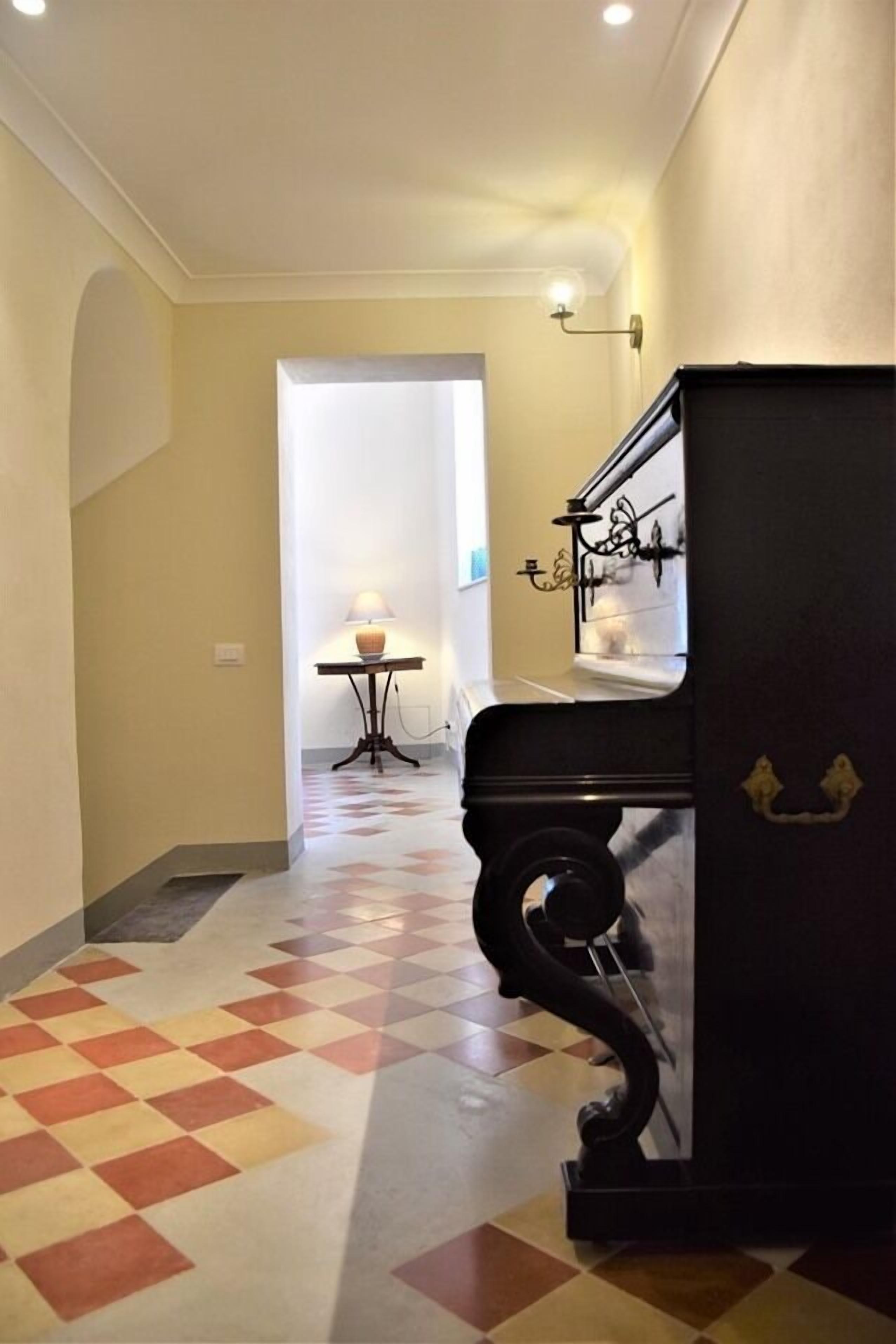Un couloir avec un sol à carreaux, un piano et une lampe au loin.